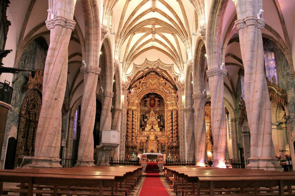 Interior de un edificio histórico con bóveda de crucería de piedra, bancos de madera alineados y una alfombra roja que conduce hacia un altar dorado, iluminado por una luz cálida y solemne.
